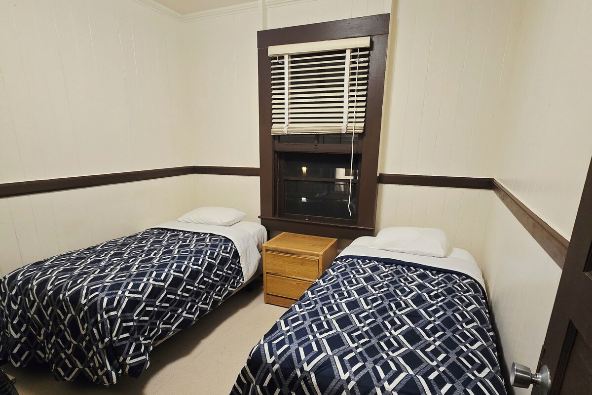 A room with two beds, a window with blinds, a wooden nightstand, and a door.