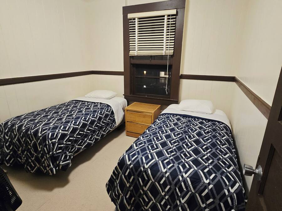 A room with two beds, a window with blinds, a wooden nightstand, and a door.