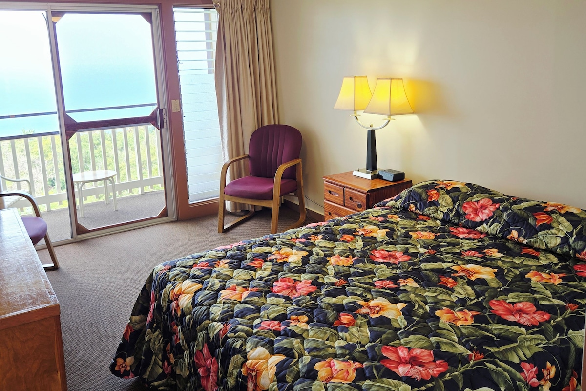 A hotel room with a bed, a chair, a desk, a lamp, and a ceiling fan.