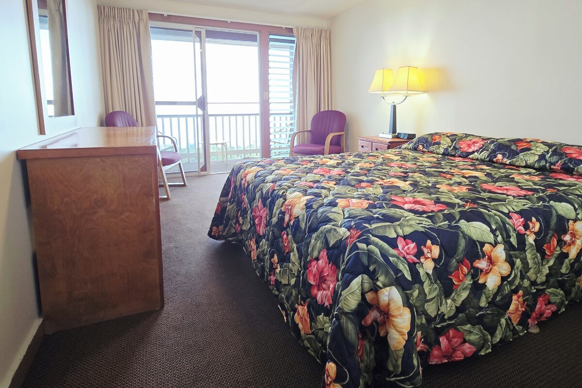 A hotel room with a bed, a desk, a chair, a lamp, and a ceiling fan.
