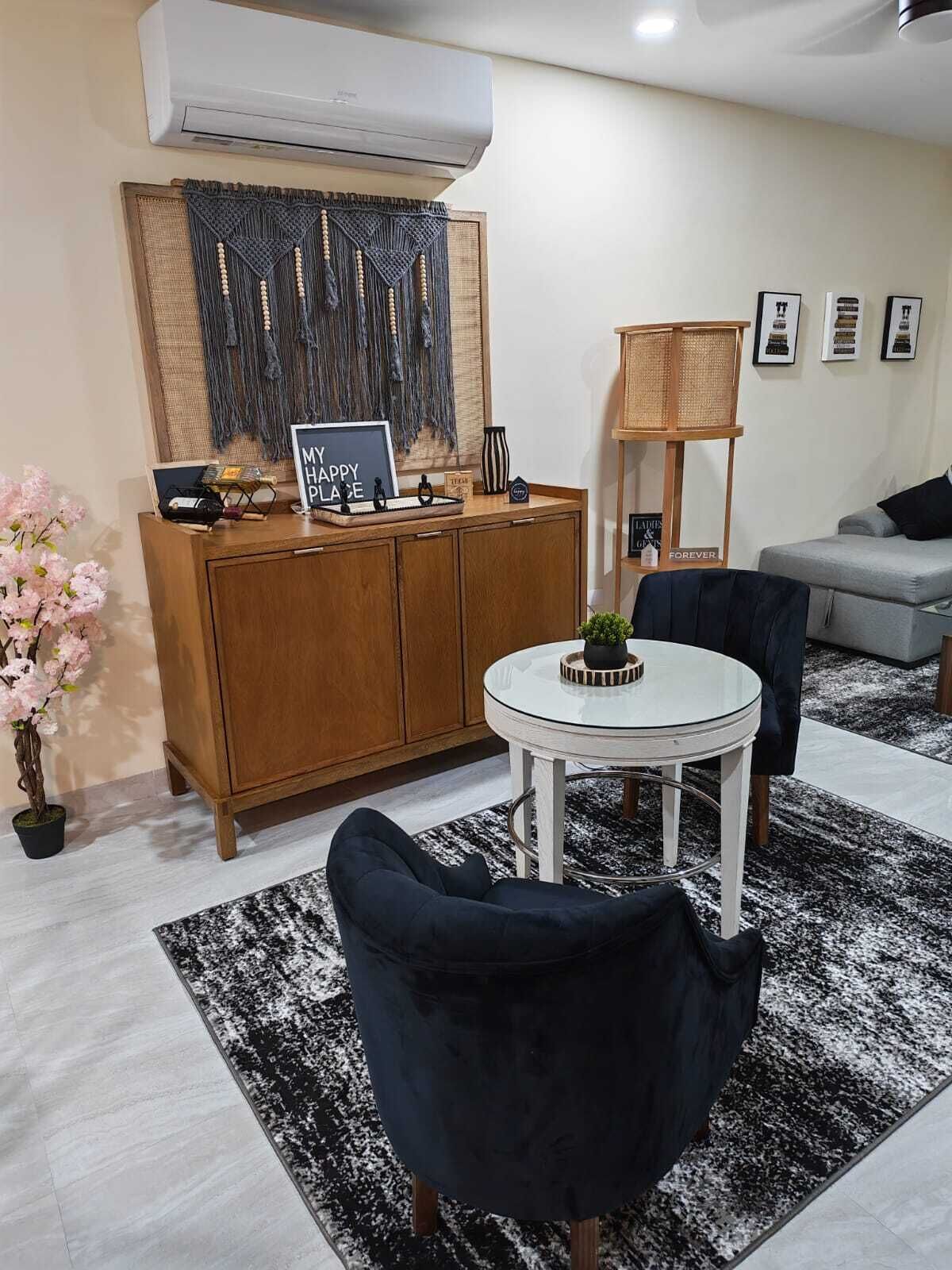 A living room with a dark blue armchair, a small white table, a wooden cabinet, and a decorative macrame wall hanging.