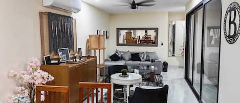 Una sala de estar moderna con un sofá, una mesa de centro y sillas. Hay un comedor con una mesa y sillas. Un espejo grande está en la pared, y hay un ventilador de techo.