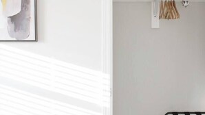 Una habitación con pared blanca, un cuadro abstracto enmarcado, un armario blanco con ropa colgada y un banco de metal negro.