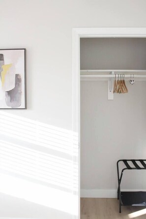 Ein Zimmer mit weißer Wand, einem gerahmten abstrakten Gemälde, einem weißen Schrank mit Kleidung und einer schwarzen Metallbank.