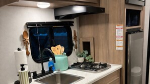 A compact kitchen with wooden cabinets, a stainless steel sink, a microwave, and a built-in oven. - Cozy Family Camper with Bunks (Fayetteville)