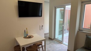 Una sala de estar con televisor de pantalla plana montado en la pared, una mesa de comedor con sillas, un sofá y un ventanal corredero que da a un espacio exterior.