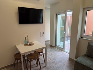 Ein Wohnzimmer mit Flachbildfernseher an der Wand, einem Esstisch mit Stühlen, einer Couch und einer Glastür, die zu einem Außenbereich führt.