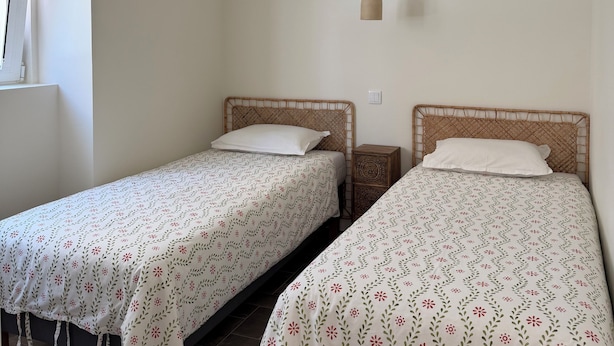 Una habitación con dos camas individuales, cada una con una colcha estampada y cabecero de madera. Hay una lámpara colgante tejida sobre las camas y una mesita de noche con un objeto decorativo.