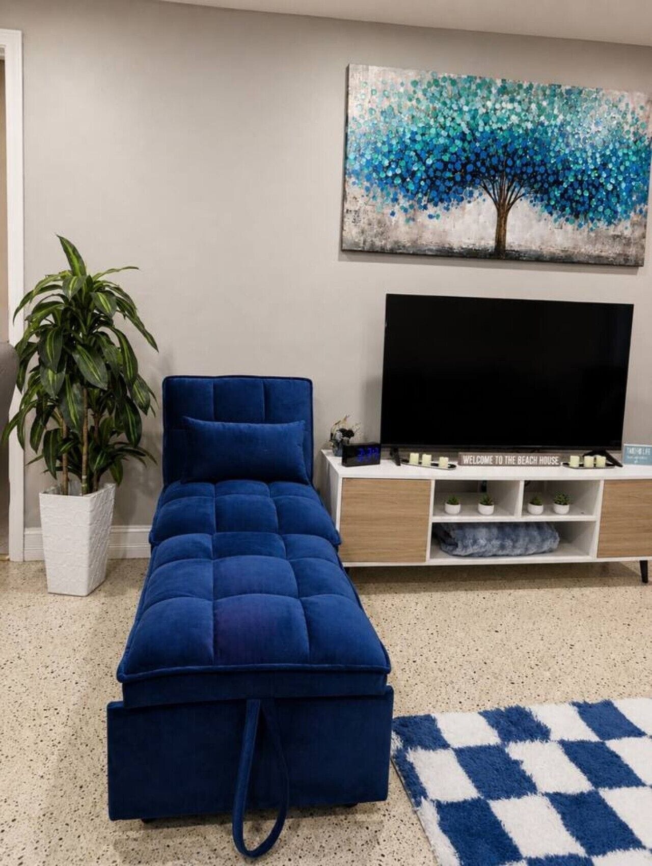 Un salon moderne avec un canapé d’angle bleu, un téléviseur à écran plat, une plante en pot et un tableau représentant un arbre.