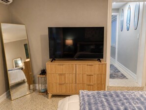 Ein Hotelzimmer mit großem Flachbildfernseher auf einer Holzkommode, einem Spiegel für den ganzen Körper und einem Bett mit gemustertem Bettüberwurf.