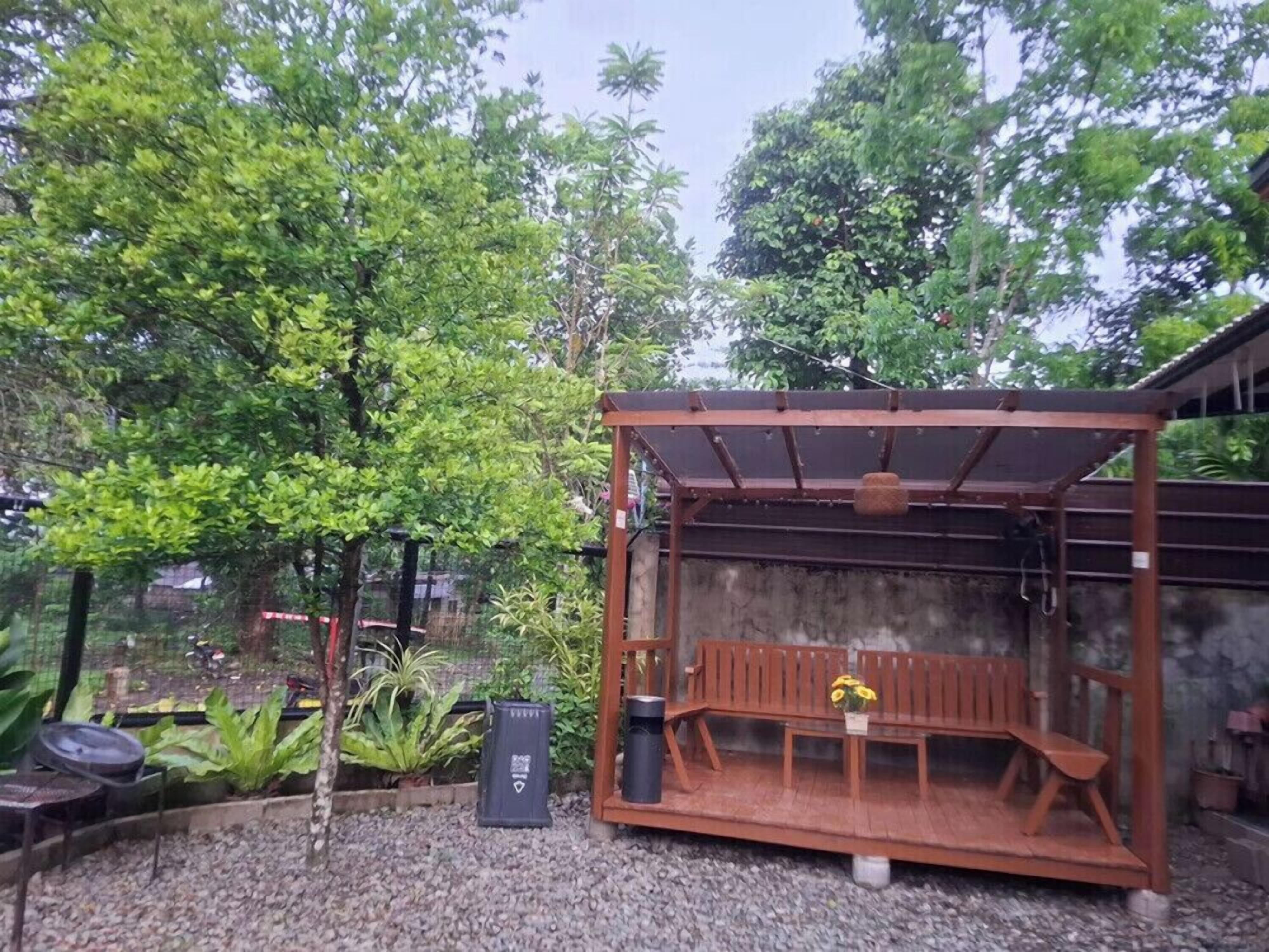 A covered outdoor seating area with a bench and table, surrounded by greenery and a concrete wall.