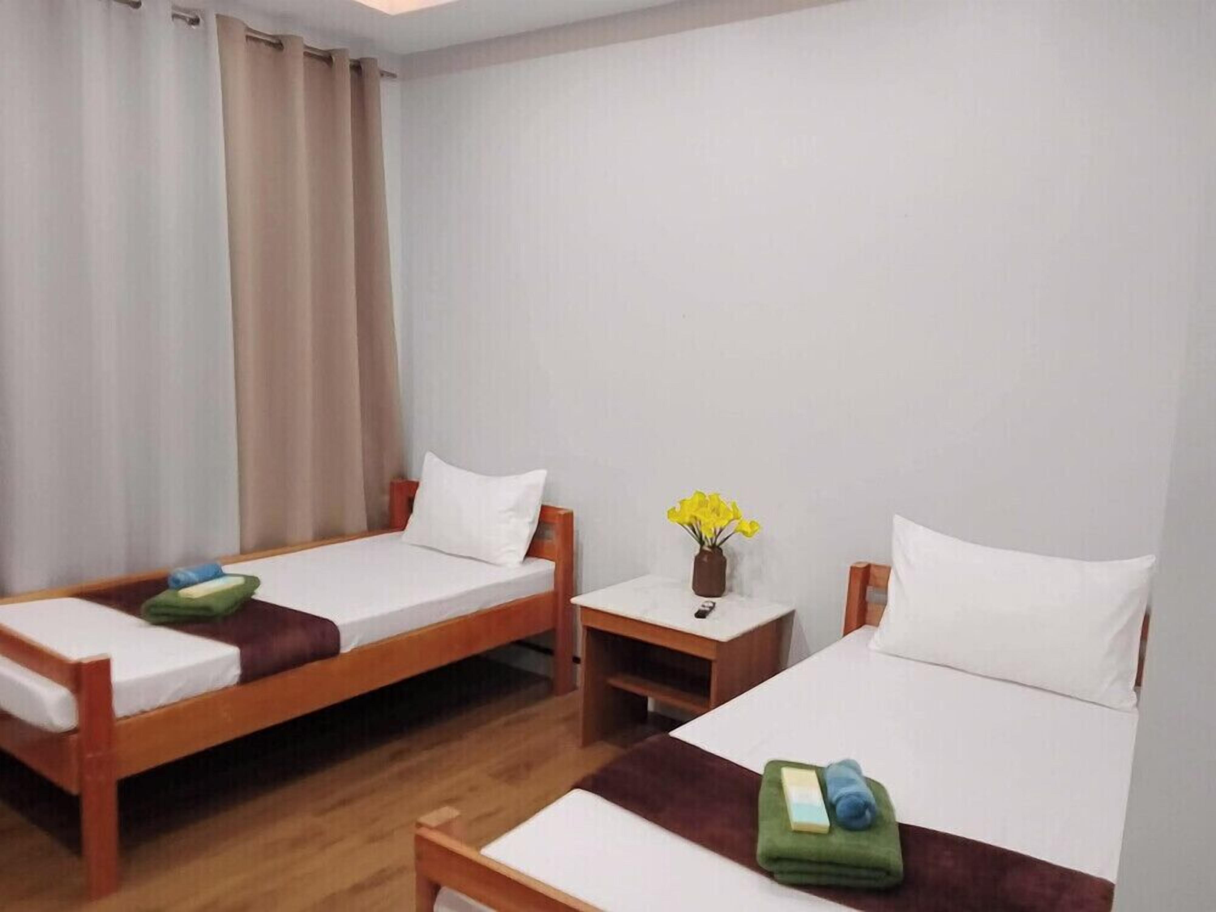 Une chambre d’hôtel avec deux lits simples, des têtes de lit en bois, du linge de lit blanc, une petite table de chevet ornée d’un vase de fleurs et un rideau à la fenêtre.