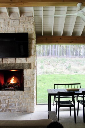 A stone fireplace with a TV mounted above it, a dining table with chairs, and a view of a grassy area with trees. - Architect-Designed Retreat on 5 Acres - Serenbe Trail Access and Detached Studio (Newnan)