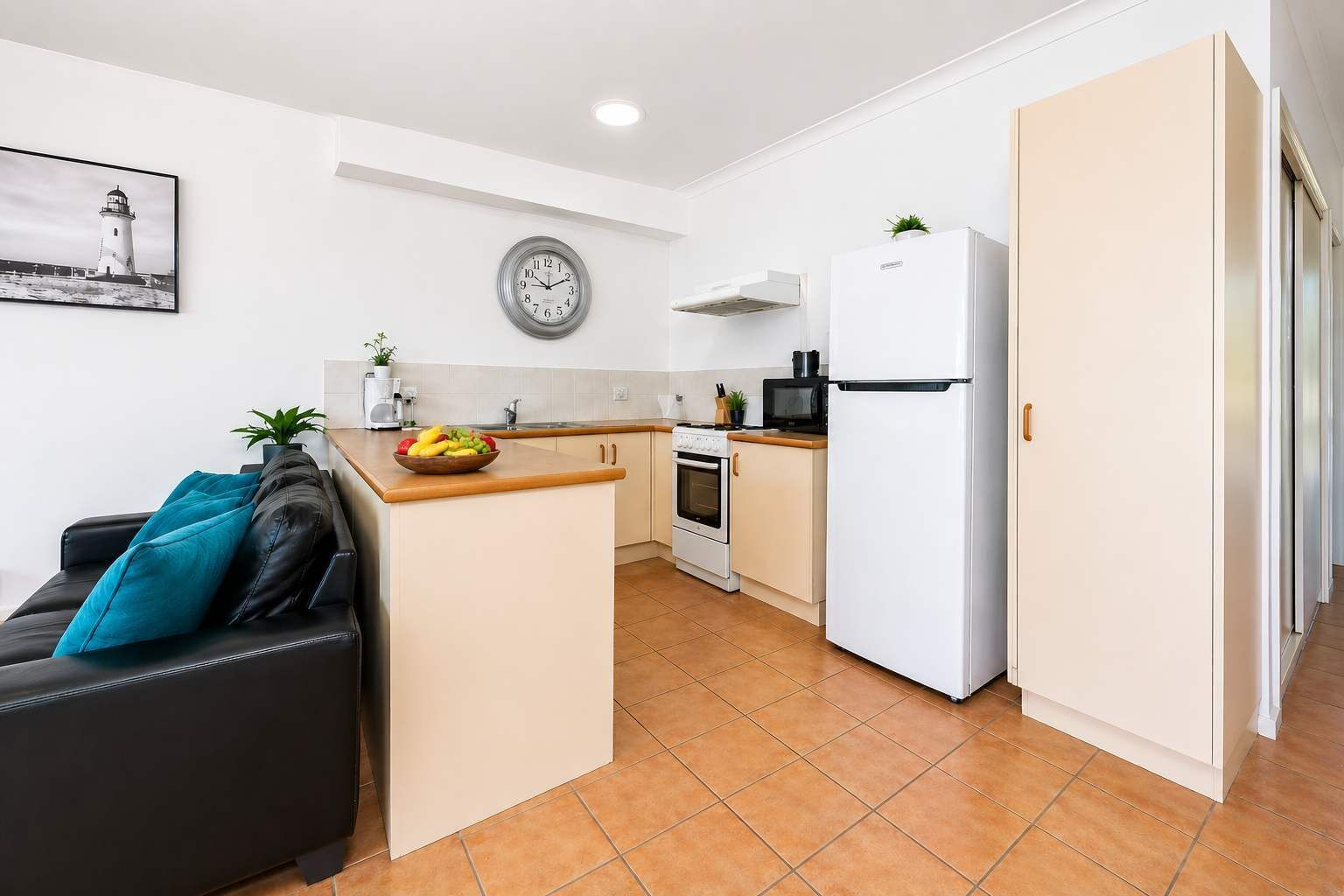 A compact kitchen with a stove, microwave, and refrigerator, a dining area with a table and chairs, and a living area with a sofa and a wall clock.