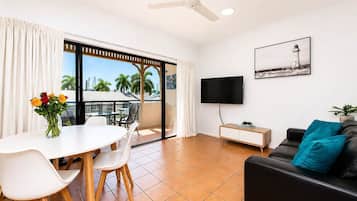 A modern living room with a dining area, a flat-screen TV mounted on the wall, and a balcony with a view of palm trees.