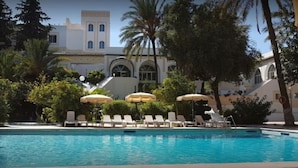 Una piscina con sillas de descanso y sombrillas, rodeada de árboles y un edificio con ventanas arqueadas.