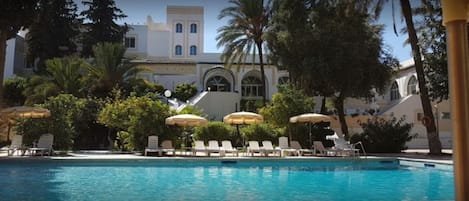 A swimming pool with lounge chairs and umbrellas, surrounded by trees and a building with arched windows.