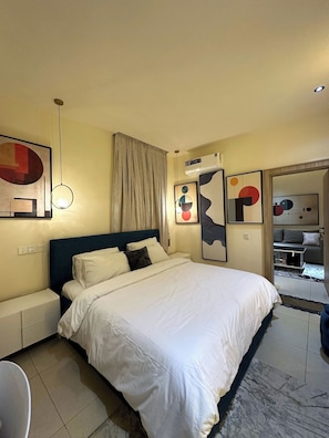 A hotel room with a bed, a bedside table, a chair, a wall-mounted air conditioner, and abstract art on the walls. - Purple Crown Hotel  (Ikeja)
