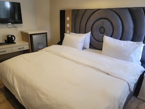 A hotel room with a large bed, a flat-screen TV mounted on the wall, a minibar, and a small desk with a coffee maker.