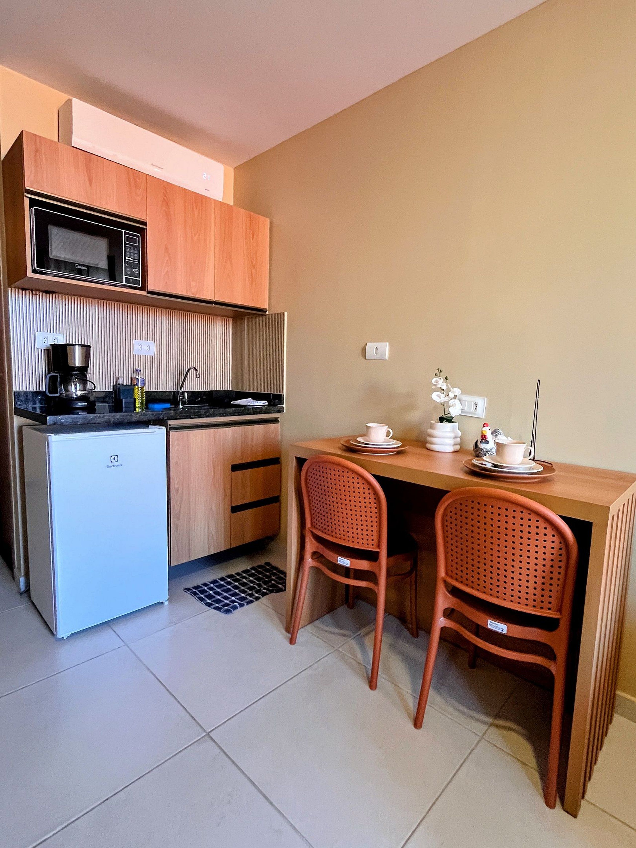 Cocina compacta con microondas, cafetera y un refrigerador pequeño. Hay una mesa de comedor con dos sillas y un pequeño adorno sobre la mesa.