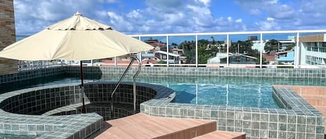 A rooftop pool area with lounge chairs, a hot tub, and a view of the cityscape.
