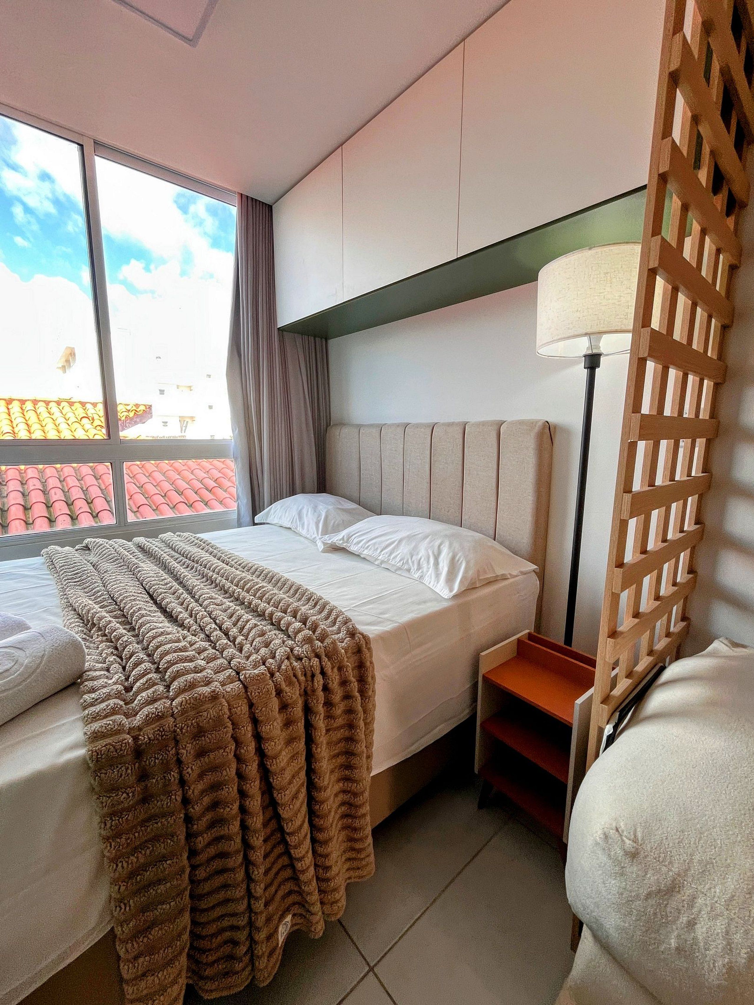 Habitación de hotel con cama, mesita de noche, lámpara, ventana con cortinas y vistas a edificios.