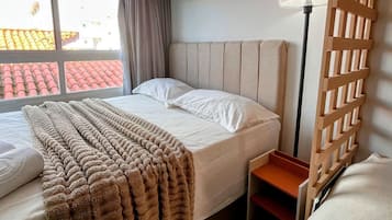Habitación de hotel con cama, mesita de noche, lámpara, ventana con cortinas y vistas a edificios.