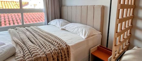 Habitación de hotel con cama, mesita de noche, lámpara, ventana con cortinas y vistas a edificios.