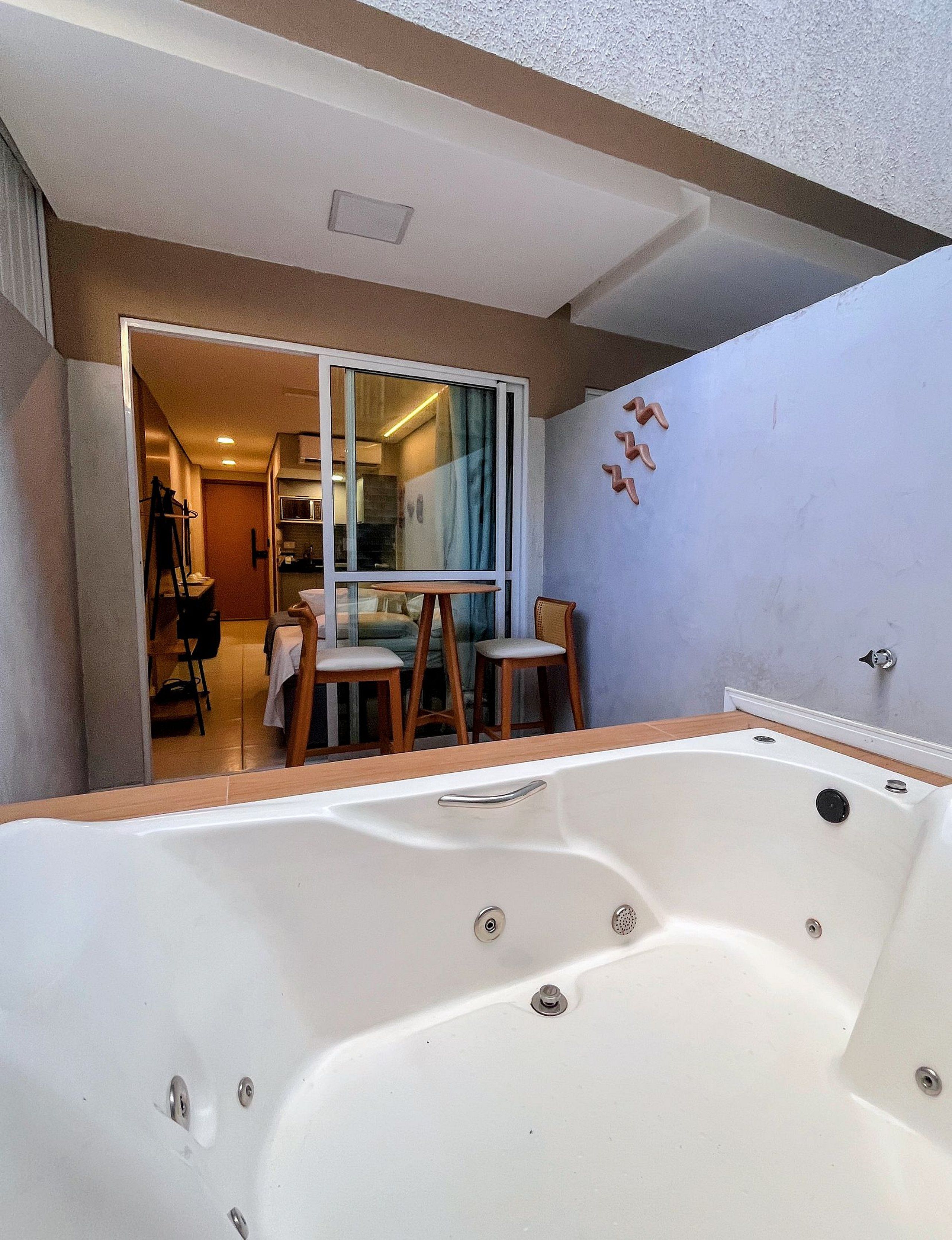 Un bagno con una vasca, una doccia e un water. Si vede una finestra con tende e una sedia sullo sfondo.