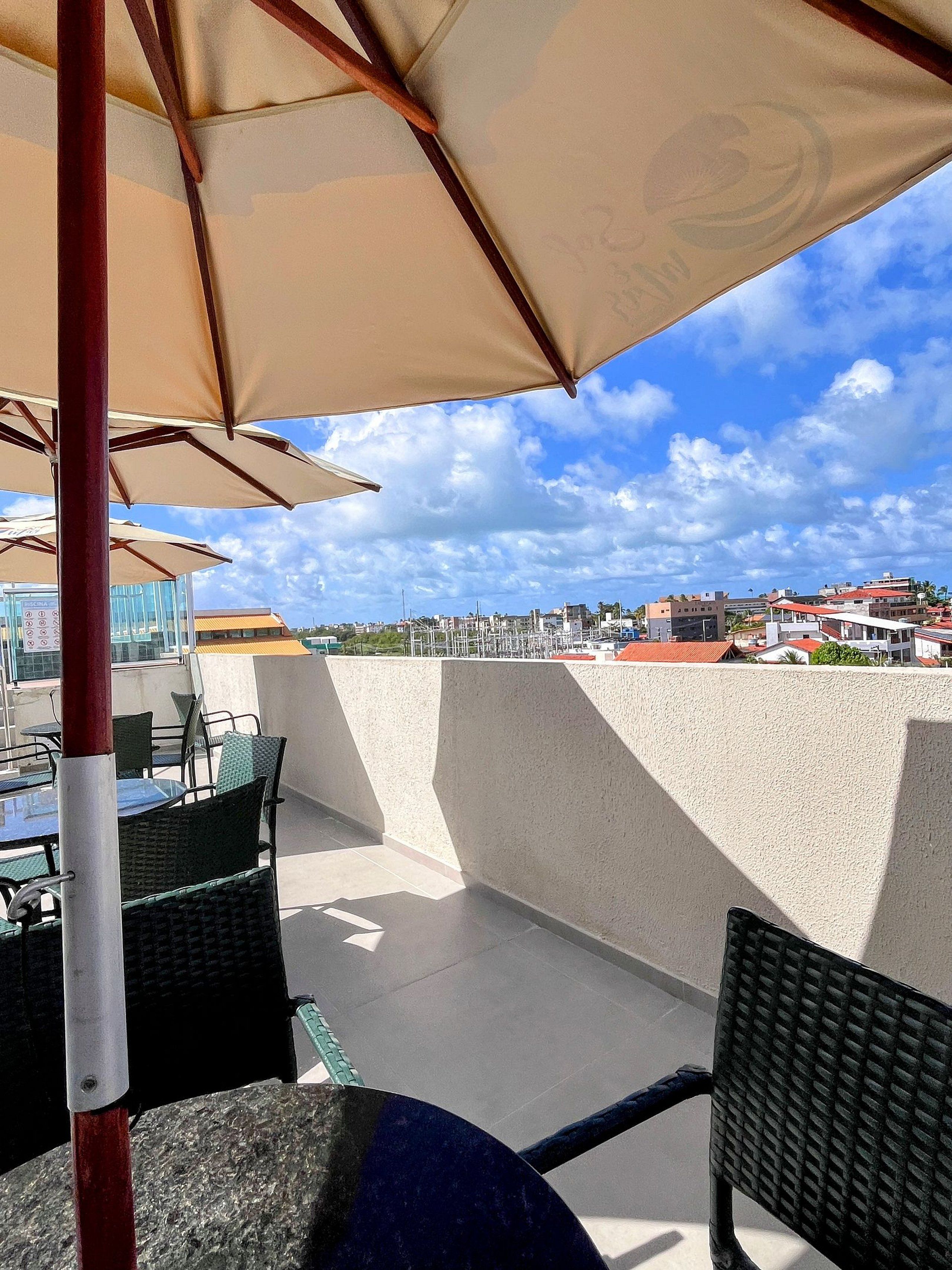An outdoor seating area with tables and chairs under umbrellas, overlooking a cityscape with buildings and a clear sky.