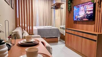 Habitación de hotel con zona de comedor, cama, televisor montado en la pared y ventana con cortinas.