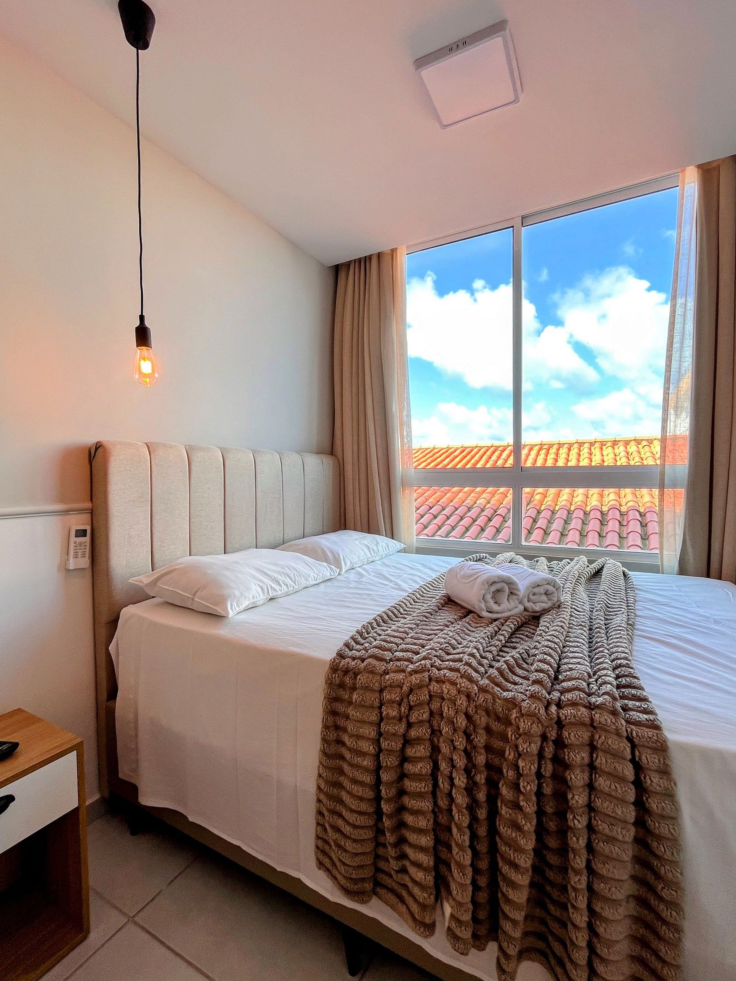 Una habitación de hotel con cama, mesita de noche, ventana con cortinas y lámpara colgante.