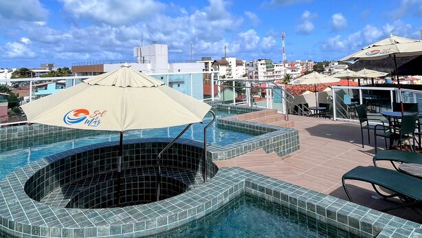 Una piscina en la azotea con borde de azulejos, una zona de estar con mesas y sillas, y vista del paisaje urbano al fondo.