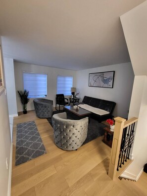 A modern living room with a sofa, two tufted ottomans, a dining table, and a staircase with a wooden railing. - Large private room in the basement | Quiet neighborhood in Aylmer | Free parking (Gatineau)
