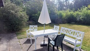 Um pátio externo com uma mesa e cadeiras brancas, um banco branco e um guarda-sol branco.