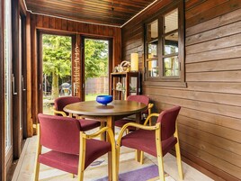 A wooden interior with a round table and four chairs, a window with a view of greenery, and a shelf with various items.