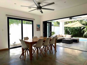 A modern dining area with a wooden table and chairs, a ceiling fan, and a view of a pool and greenery outside. - Quality Home in Perfect Location Close to Beach & Restaurants (Sámara)