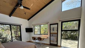 Een slaapkamer met een bed, een bureau en een stoel, grote ramen en een plafondventilator.