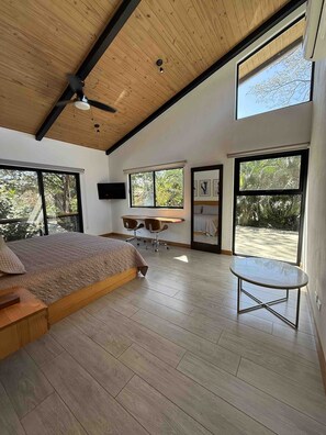 Ein Schlafzimmer mit einem Bett, einem Schreibtisch und einem Sessel, großen Fenstern und einem Deckenventilator.