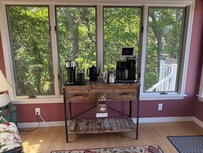 Een houten tafel met koffiezetapparatuur en een bordje met de tekst 'Coffee Happens Here' voor een raam met uitzicht op bomen.