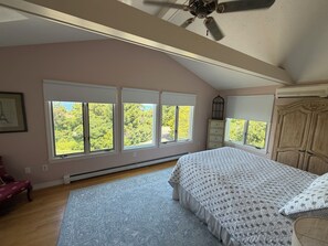 Une chambre à coucher comprenant un lit, un ventilateur de plafond, une fenêtre donnant sur des arbres, une commode et une chaise.