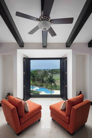 A room with a ceiling fan, two orange armchairs, a large window with black shutters, and a view of a swimming pool and greenery. - Ocean & Golf View Penthouse | Private Chef & Cart (Punta de Mita)