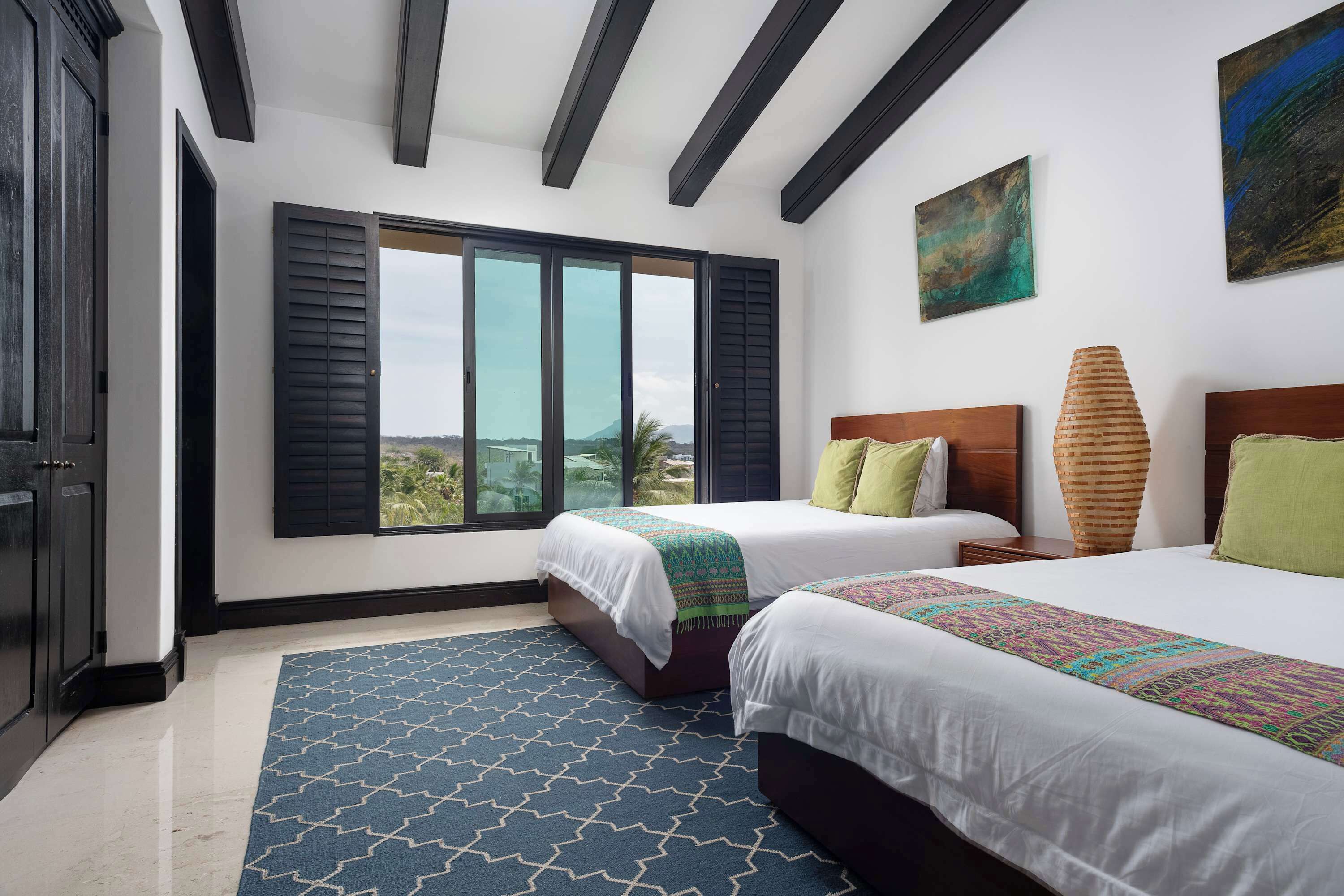 A hotel room with two beds, a large window with shutters, a painting on the wall, and a patterned rug.