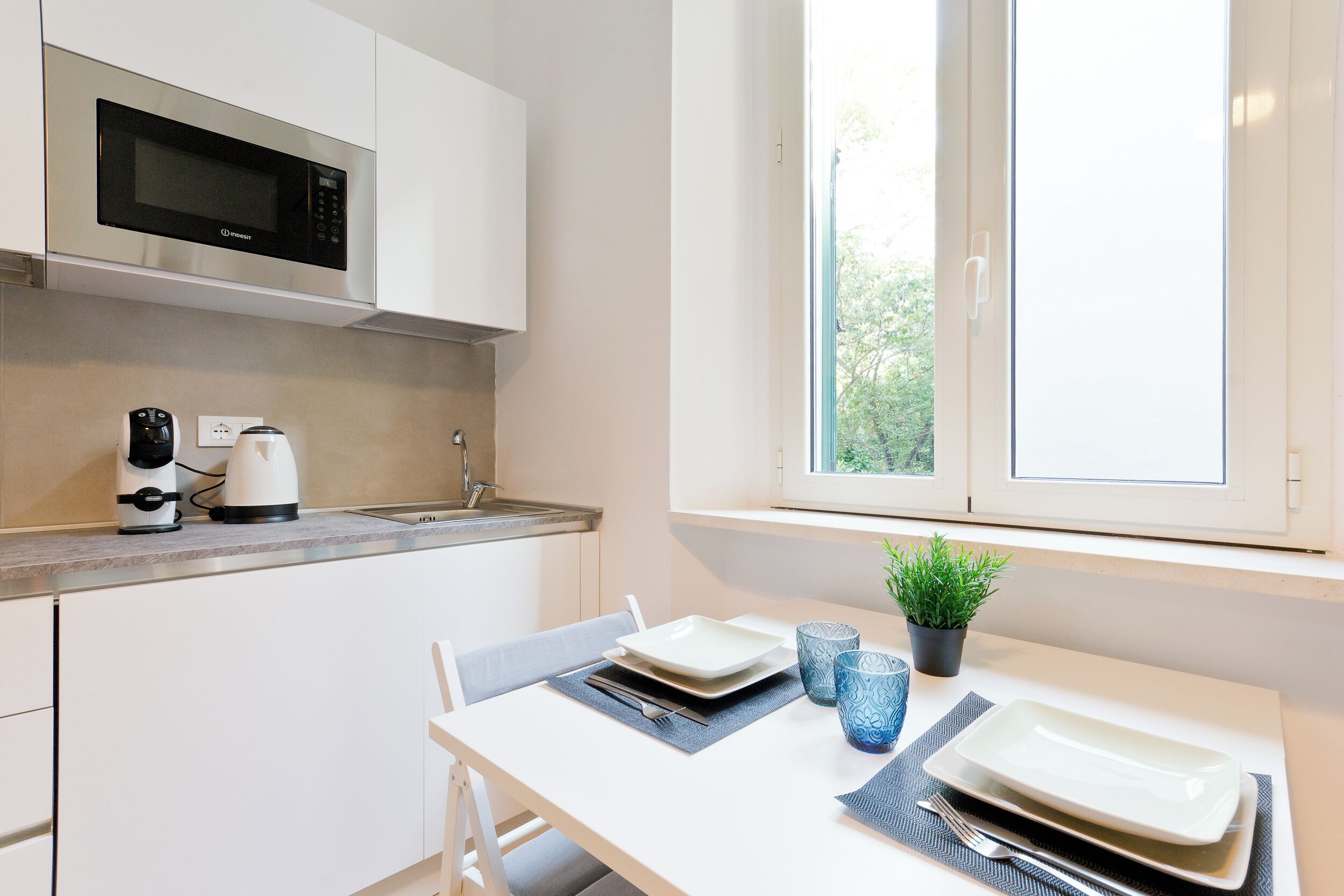 A modern kitchen with white cabinetry, a built-in microwave, a coffee maker, and a window with a view of greenery.