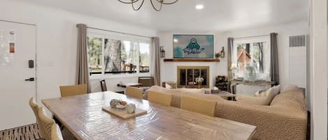A dining area with a large wooden table, cushioned seating, a chandelier, and a television mounted on the wall.