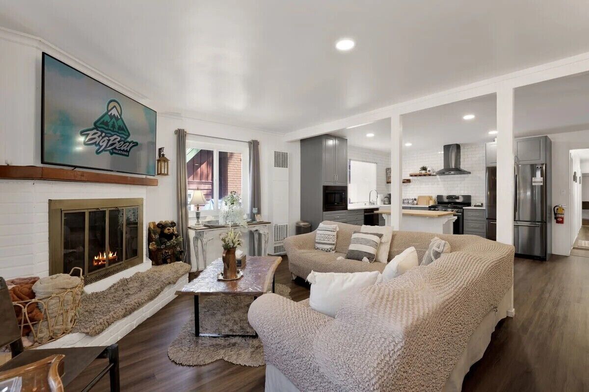 A spacious living area with a fireplace, a television mounted on the wall, and a kitchen in the background.