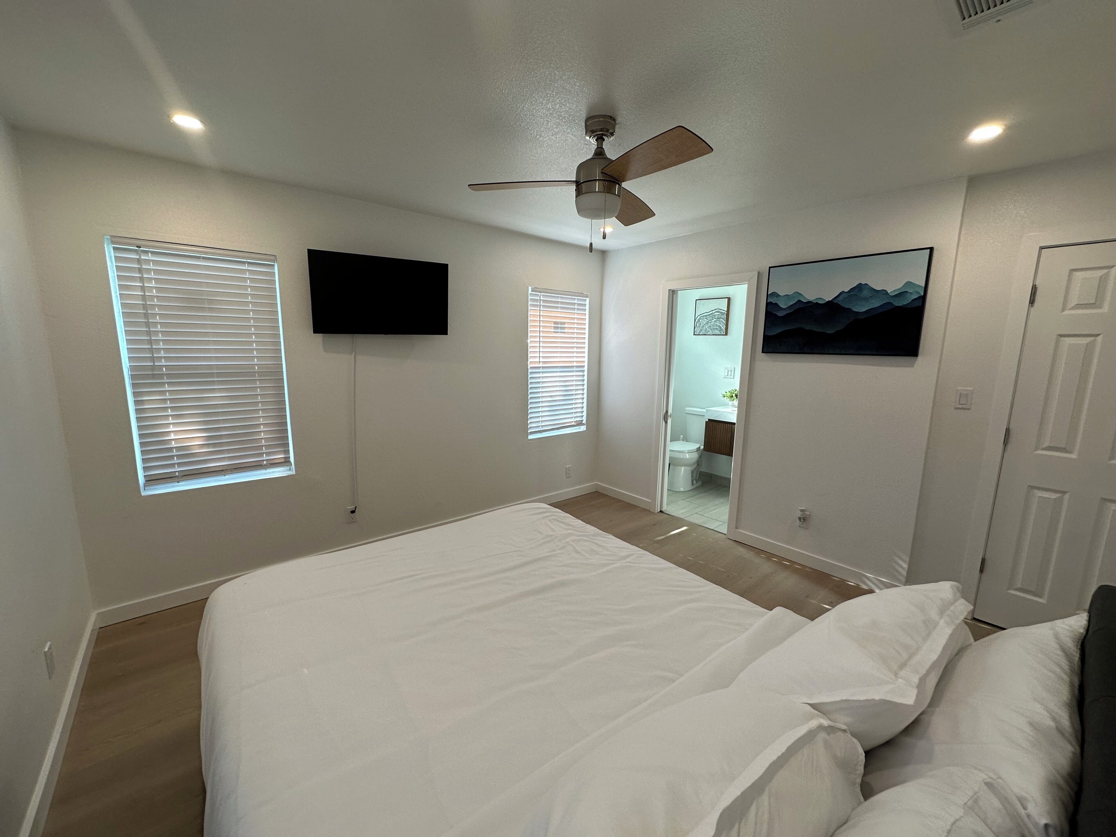 Un dormitorio con cama, ventilador de techo, televisor montado y puerta que da a un baño.
