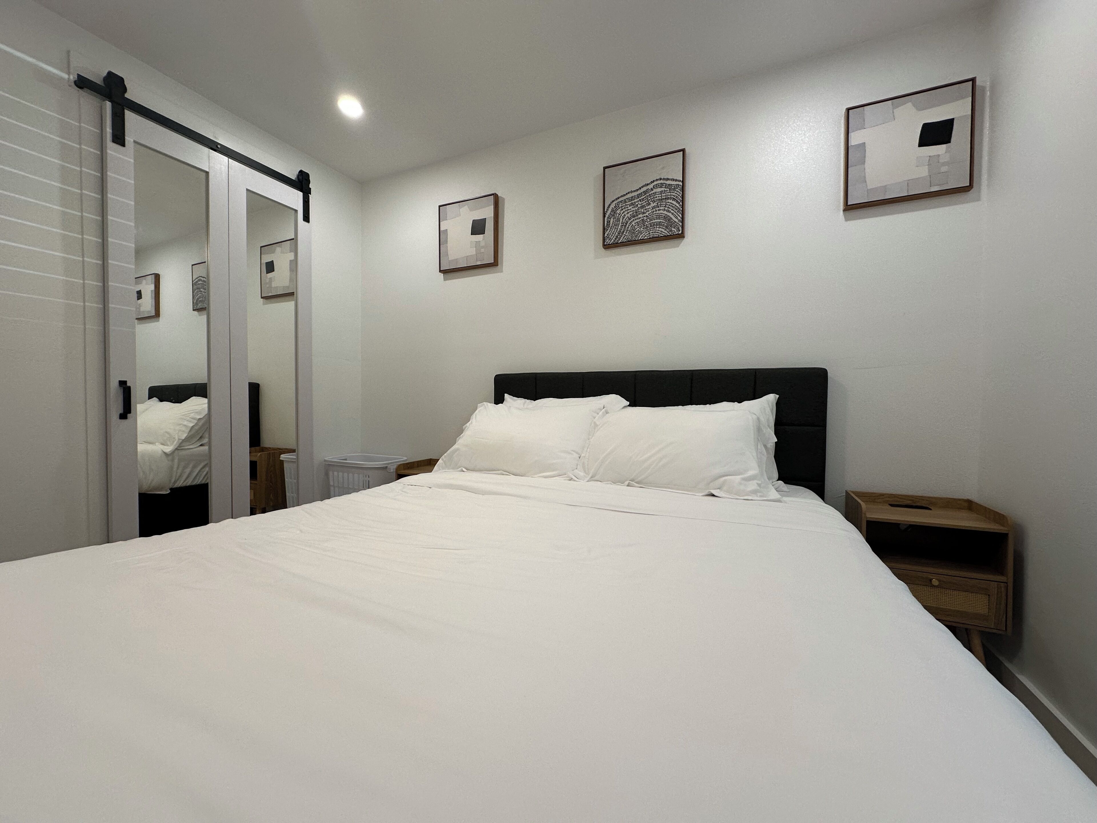 A neatly made bed with white linens in a room with white walls, a black headboard, and two bedside tables with wooden nightstands.