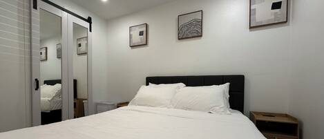 A neatly made bed with white linens in a room with white walls, a black headboard, and two bedside tables with wooden nightstands.