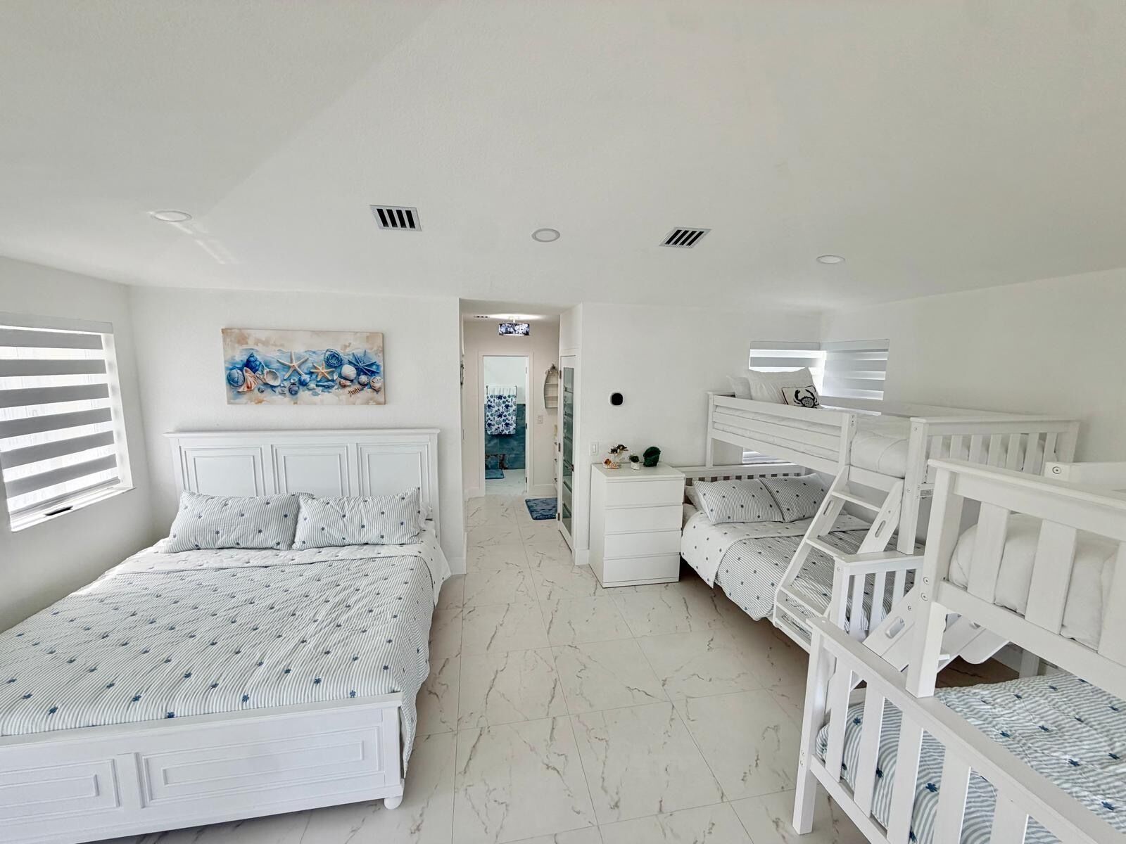 A bedroom with a large bed, a smaller bed, and a crib. There is a painting on the wall and a window with blinds.
